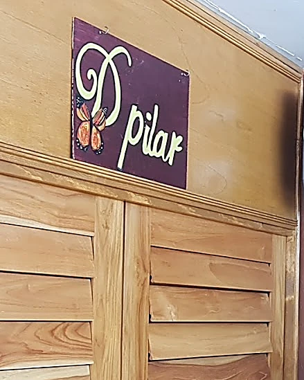 Letrero de D'Pilar: placa morada con el nombre en letra verde y una mariposa monarca naranja, sobre el muro de persianas de teca