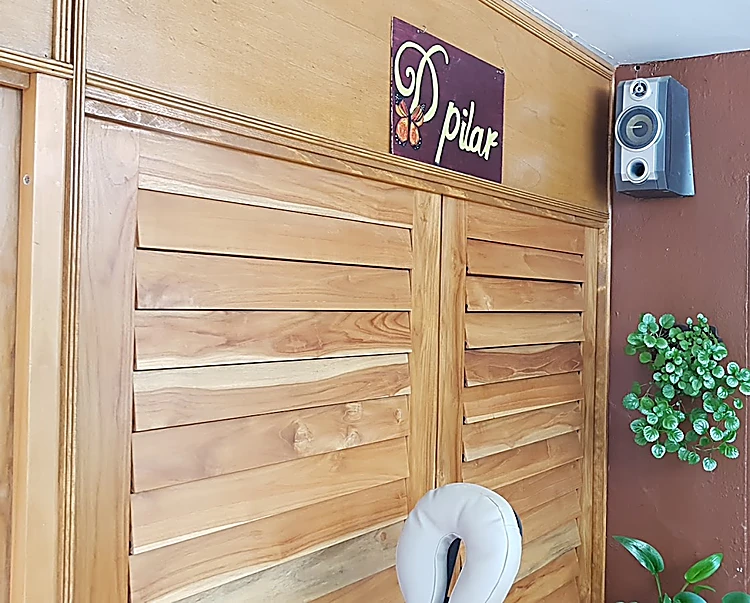 Recepción de D'Pilar: muro de persianas de teca, el letrero morado con el nombre en letra verde y una mariposa monarca, una bocina y una planta colgante